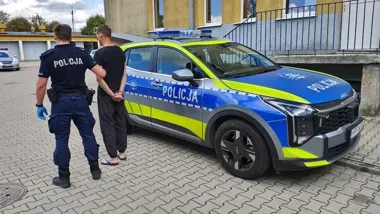 Policjant w mundurze prowadzi zatrzymanego mężczyznę do policyjnego radiowozu Kia zaparkowanego przed budynkiem.